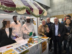 L'image montre le Maire de Bois Colombre, Yves Reveillon, au stand de miel. Il ets accompagné de ses équipe. L'enseignante et ses élèves sont derrière le comptoir du stand