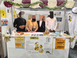 L'image montre une enseignante et 3 élèves derrière le stand de vente de miel. Des affiches pédagogiques et des tenues d'apiculteurs ont été installés en décoration