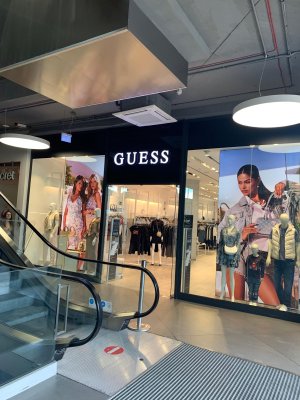 Cette image montre l'entrée du magasin Guess par l'intérieur du centre commercial. Devant il y a un escalator.