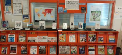 Cette image montre des livres et des périodiques exposés sur des présentoirs orange. Derrière sont affichées les affiches de la SPME 2025 et la carte de la liberté de la presse de Reporters sans frontière