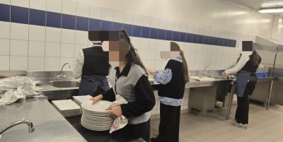 Cette image représente4 élèves dans une cuisine professionnelle. Ils s'affairent autour de grands plan de travail en intox. Au premier plan, une élève porte une pile de grandes assiettes.
