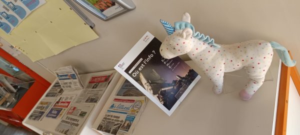 Cette image montre la peluche licorne prénommée Hermione entrain de lire le dossier du CLEMI de la SPME 2025 sur le thème "où est l'info ?". De journaux sont exposés plus loin sur la table.